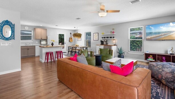 A modern living room with a brown leather sofa, a dining area with a table and chairs, a kitchen with wooden cabinets, and a large window with a beach view.