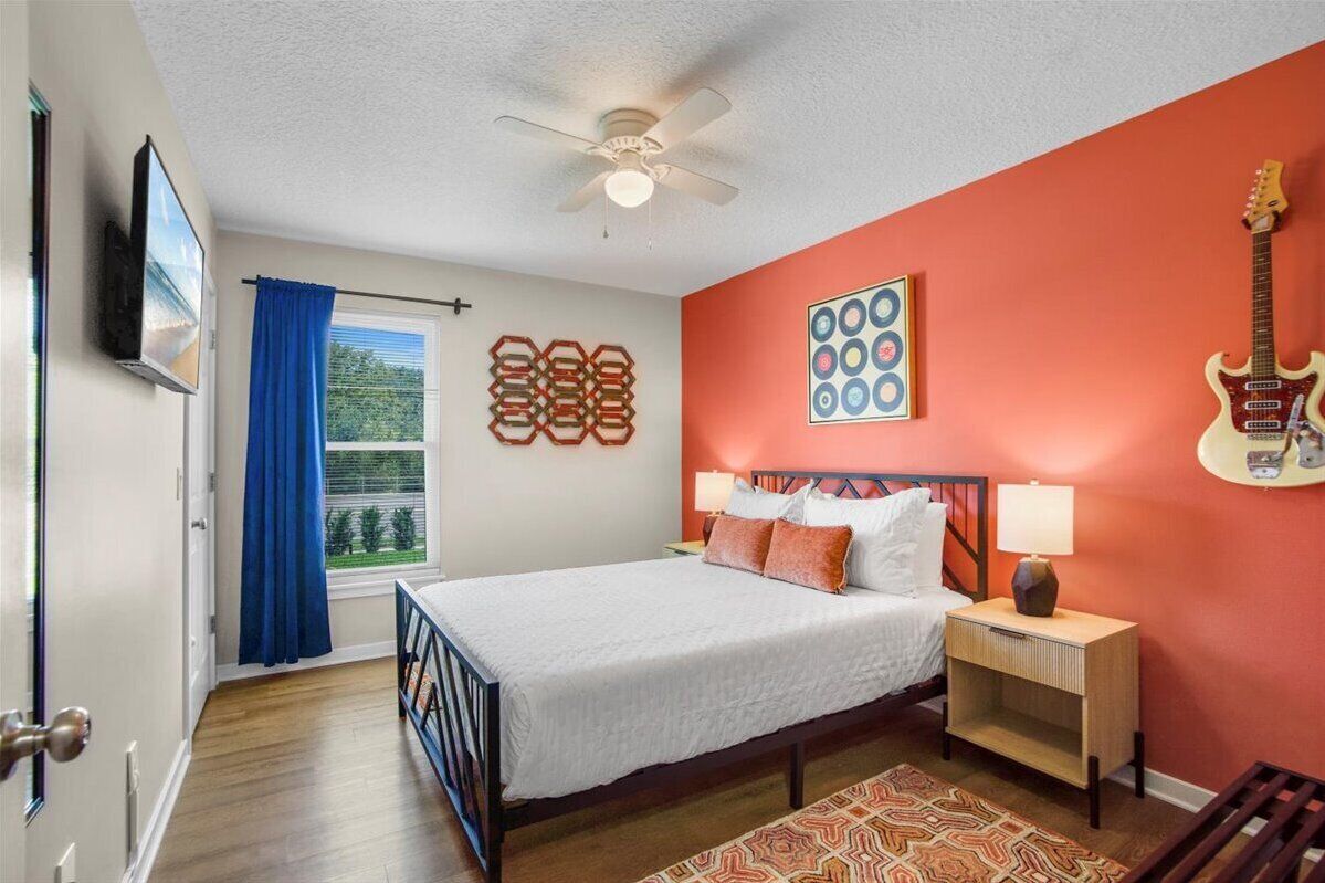 A bedroom with a bed, a ceiling fan, a window with blue curtains, and a guitar on the wall.