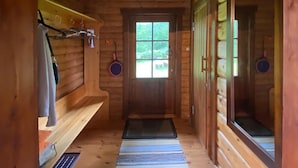 L’intérieur d’une cabane en bois, avec une porte centrale, une petite fenêtre et un banc en bois le long du mur gauche.