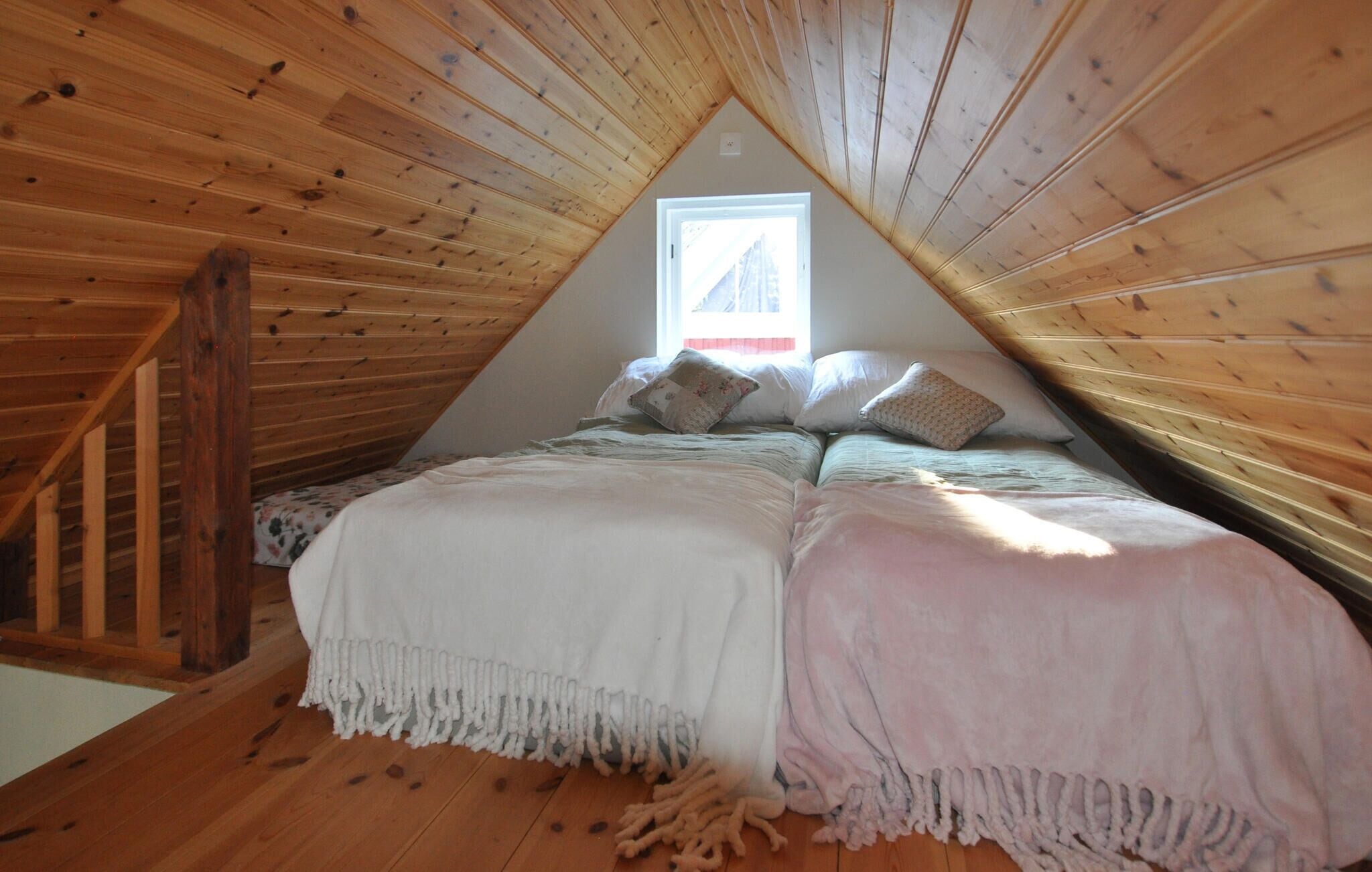 A cozy attic bedroom with a sloped ceiling, wooden beams, a window, and a bed with pillows and blankets.
