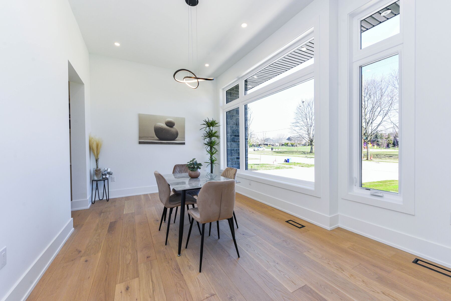 A dining area with a wooden floor, a table with chairs, a hanging light fixture, and a view of a park through large windows.
