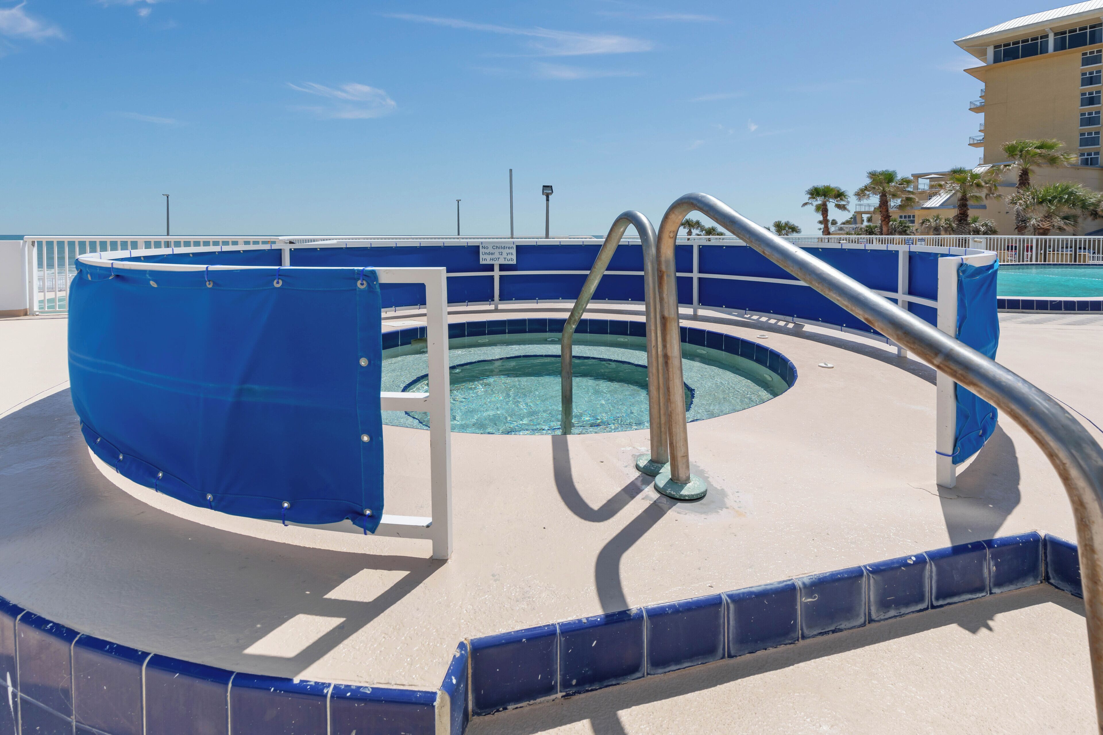 Une piscine circulaire, recouverte d’une bâche bleue et dotée d’un escalier en métal menant à l’intérieur, est entourée d’une rambarde blanche et d’une bordure bleue en carrelage.