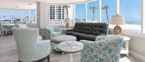 Un vaste espace de vie avec une grande fenêtre donnant sur une plage, aménagé avec un canapé, des fauteuils et une petite table ronde.