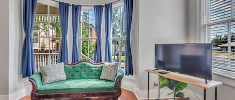 A room with a green velvet sofa, a flat-screen TV on a wooden stand, and large windows with blue curtains.