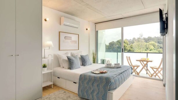 A modern bedroom with a large bed, bedside tables, a desk, and a balcony with a view of greenery. - Balcony 2BRs with Pool & Parking by HostWise (Vila Nova de Gaia)