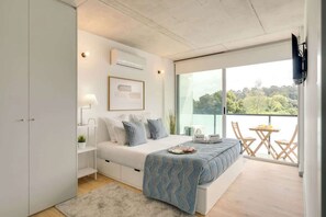 A modern bedroom with a large bed, bedside tables, a desk, and a balcony with a view of greenery.