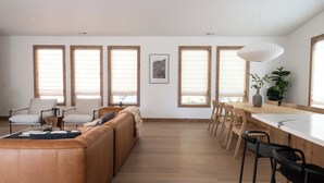 A spacious living area with a large sofa, dining table, and chairs. There are multiple windows with blinds, a pendant light, and a framed artwork on the wall. - Newly Built/Furnished Quiet Lux Ski-In/Out Hot Tub (Government Camp)