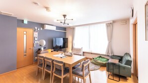 Ein modernes Wohnzimmer mit einem Holzesstisch, grauen Stühlen, einem grünen Sofa und einem Fernseher.