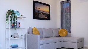 Una sala moderna con un sofá seccional gris, una fotografía enmarco de una playa, un estante blanco con objetos decorativos, y una ventana con cortinas traslúcidas.