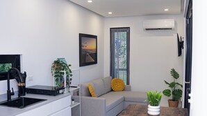 Un espacio habitable moderno con un sofá gris, una pequeña mesa de comedor, una zona de cocina con fregadero y un ventanal con cortinas.