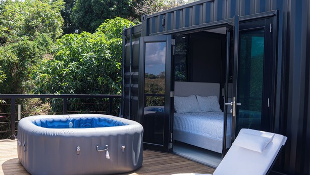 Un dormitorio moderno al aire libre con una cama, una silla de descanso y un jacuzzi inflable en una terraza de madera.