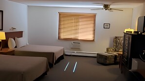 Une chambre d’hôtel avec deux lits, un bureau avec une lampe, un ventilateur de plafond, une fenêtre avec des stores et un téléviseur.