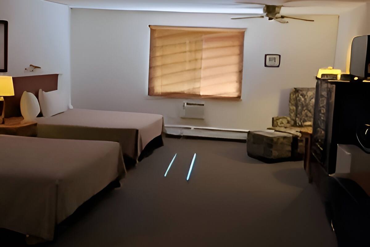 Een hotelkamer met twee bedden, een bureau met lamp, een plafondventilator, een raam met jaloezieën en een televisie.