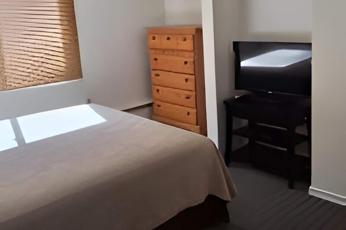 Een slaapkamer met een bed, een houten dressoir, een televisie en een raam met jaloezieën.