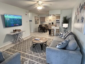 A living room with a gray sofa, a coffee table, and a TV displaying a sports game. - Spacious Beachside Condo in Cocoa Beach, Florida! 125 Feet to Beach! (Cocoa Beach)