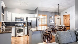 Una cocina moderna con electrodomésticos de acero inoxidable, un comedor con mesa de madera y taburetes de bar, y una zona de estar acogedora con un sofá gris.