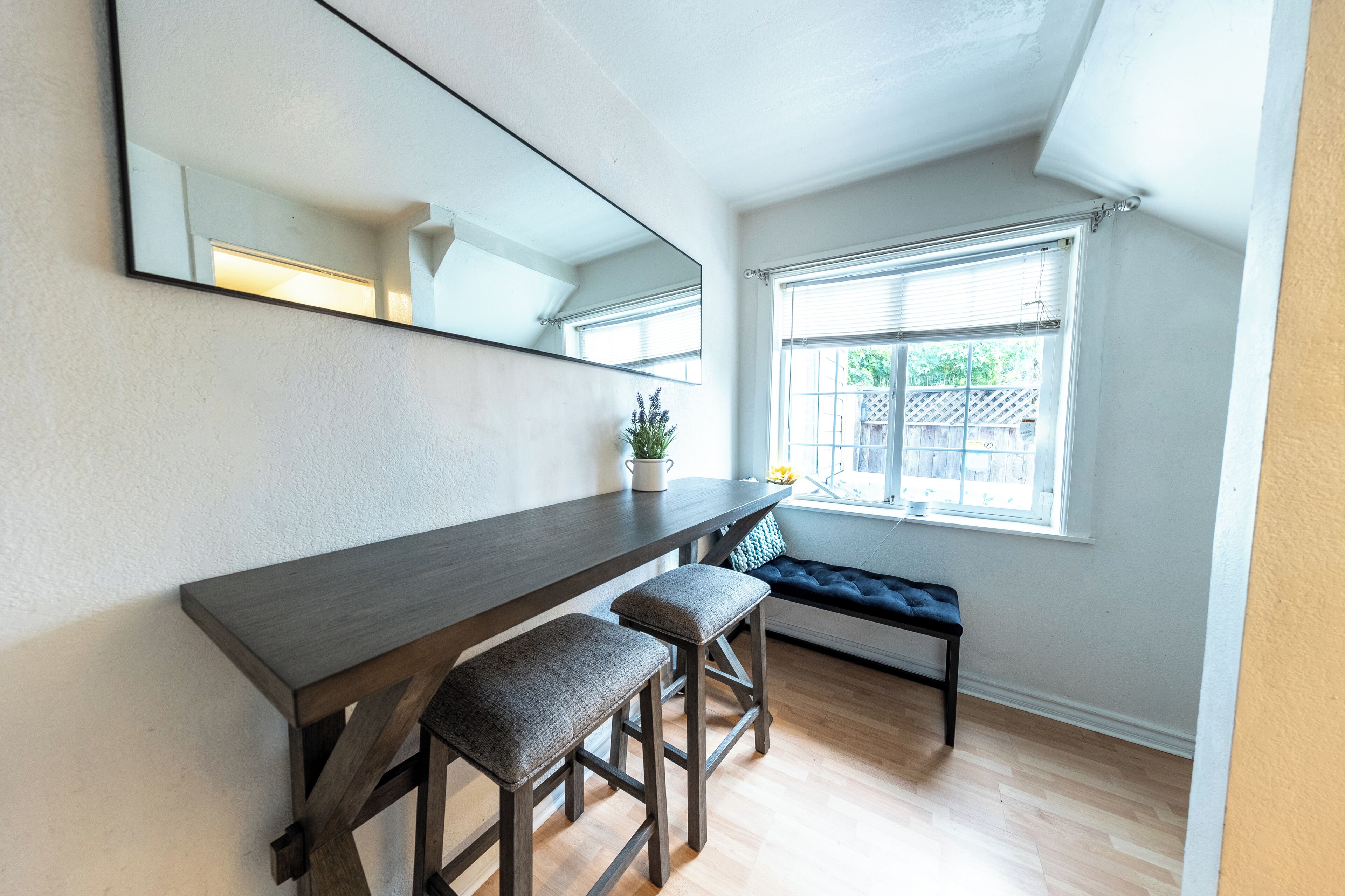 A modern room with a dark wooden desk, two cushioned stools, a large mirror, and a window with blinds.