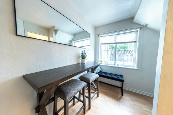 A modern room with a dark wooden desk, two cushioned stools, a large mirror, and a window with blinds.