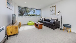 A modern living room with a large window, a leather sofa, a wooden coffee table, a green bean bag, a floor lamp, and a TV on a wooden cabinet.
