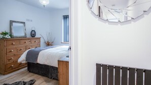 A bedroom with a wooden dresser, a bed with a grey headboard, a decorative mirror, and a modern light fixture.