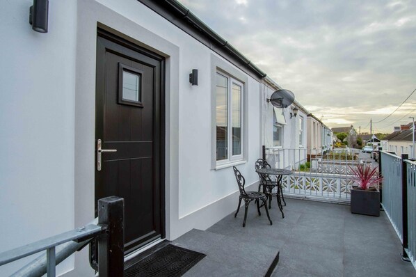 A white building with a black door, a small patio with a table and chairs, and a white fence.