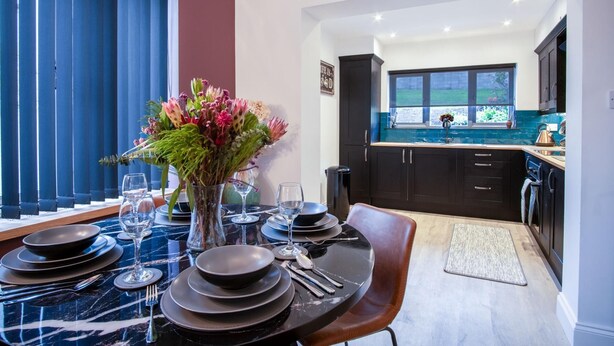 A dining area with a table set for six, a vase of flowers, and a view into the kitchen.