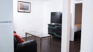 Ett kompakt vardagsrum med ett bord med glasskiva, en svart lädersoffa, en tv-apparat placerad på en svart och turkosfärgad byrå och en inramad tavla på väggen.