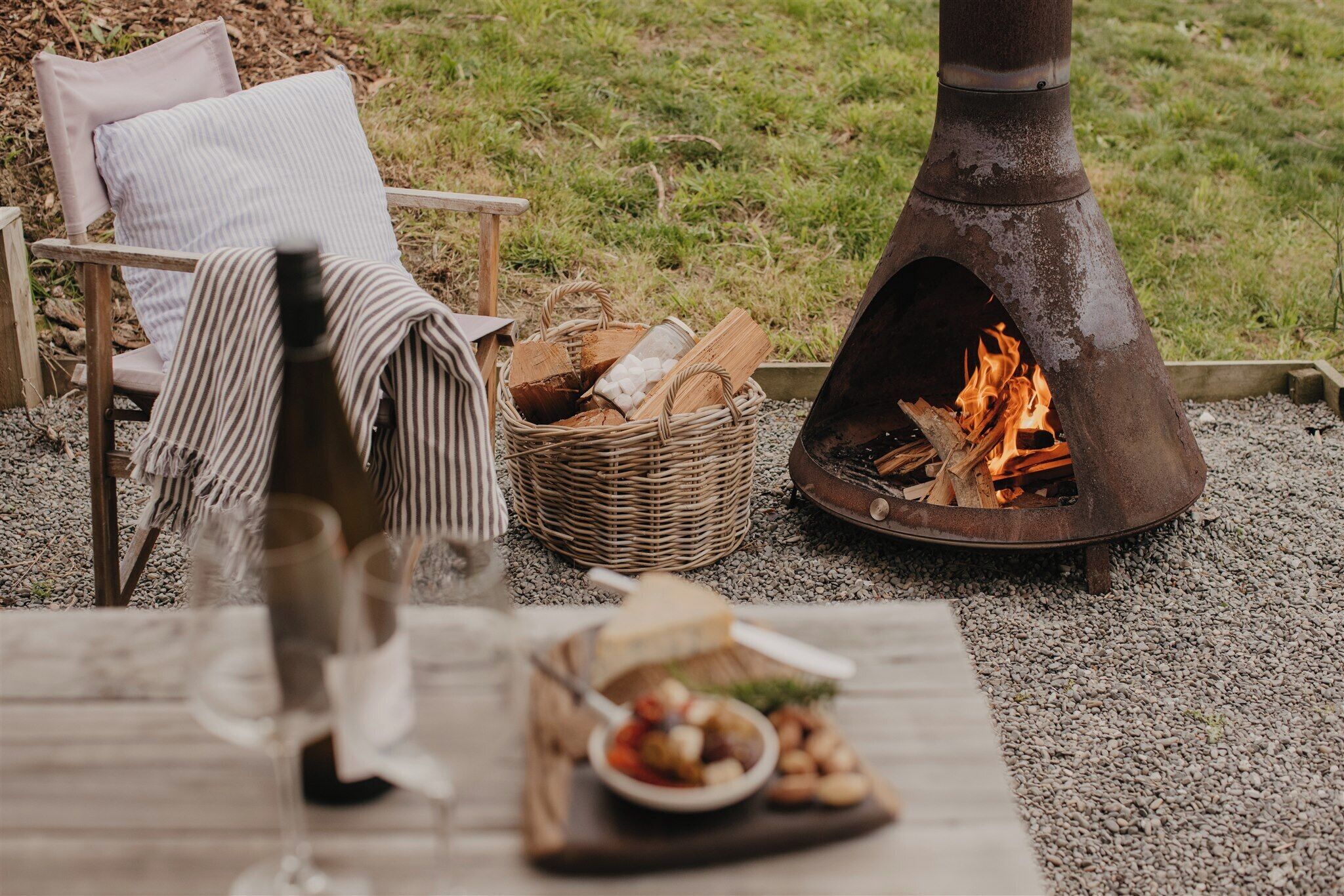 A cozy outdoor seating area with a wicker basket, a chair with a striped cushion, and a table with a plate of food and a glass.
