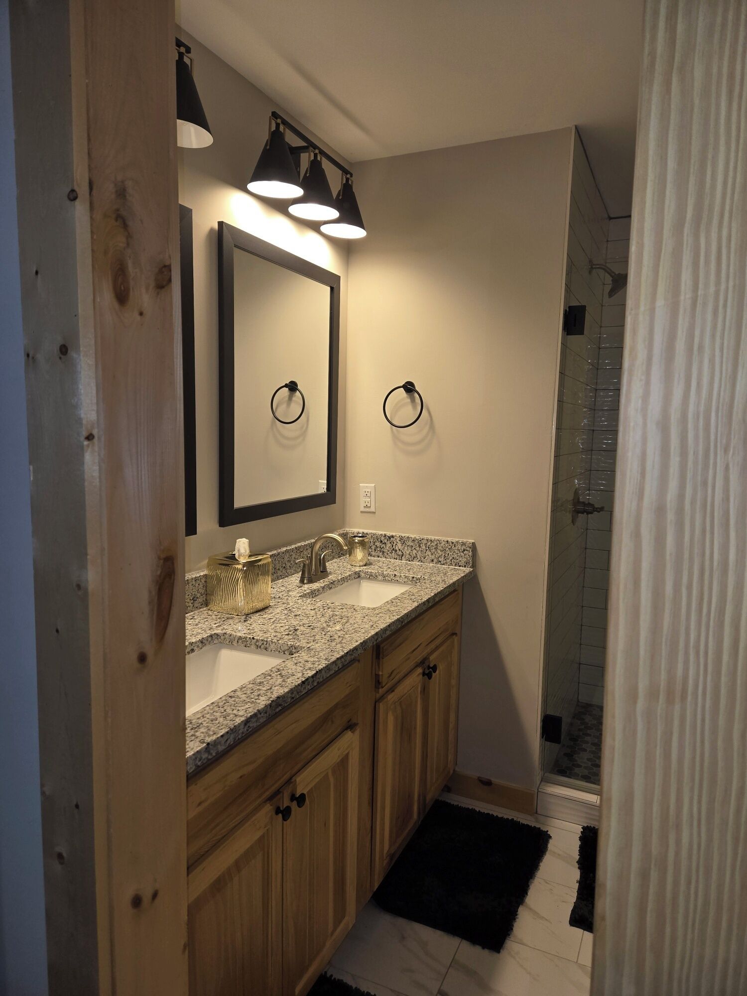 A modern bathroom with a granite countertop sink, wooden cabinets, and a glass shower enclosure.