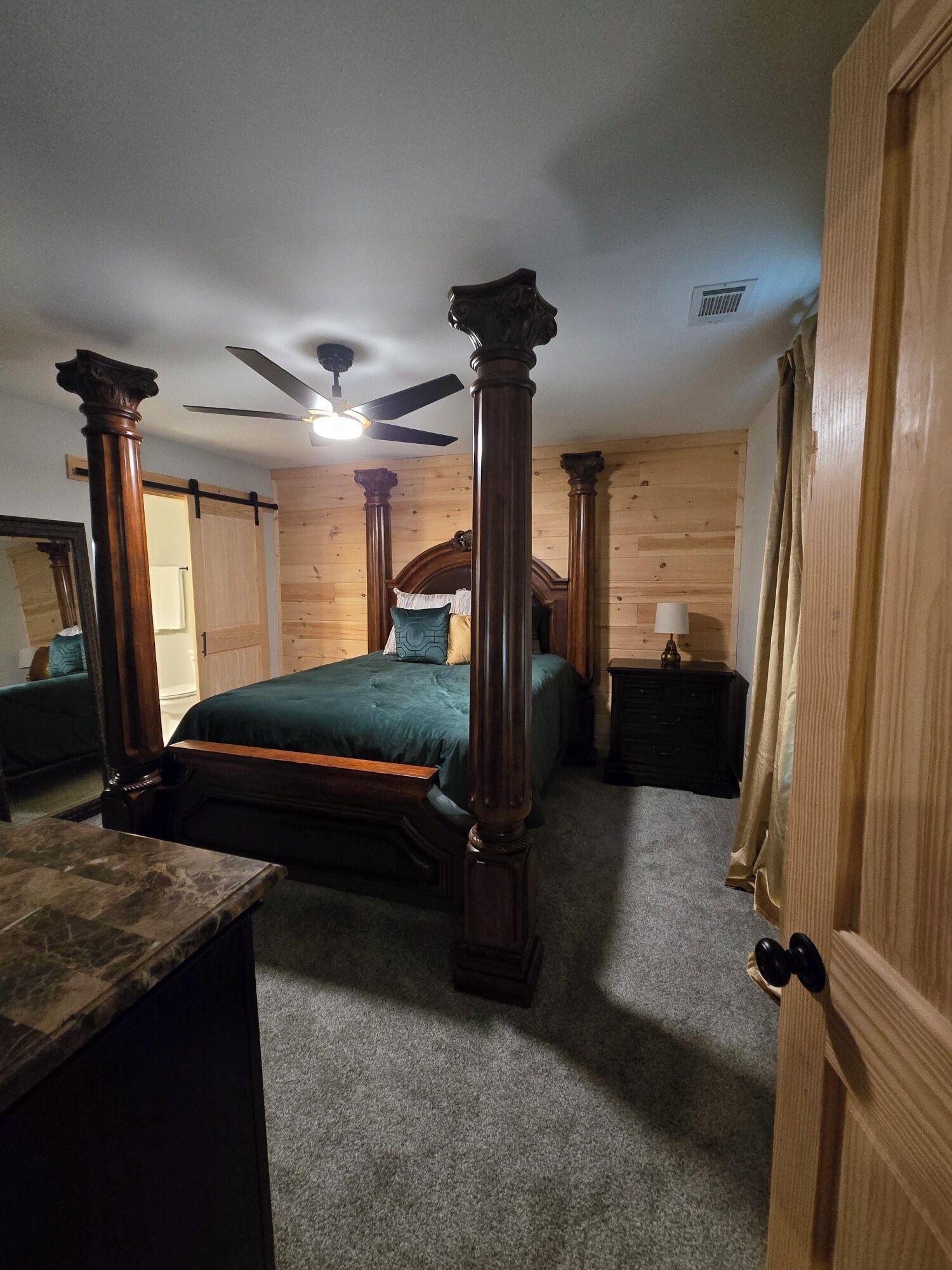 Een slaapkamer met een hemelbed, houten wanden, een plafondventilator en een nachtkastje met een lamp.