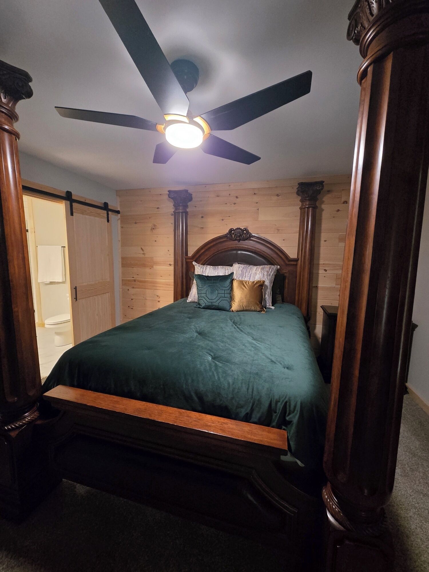 A bedroom with a bed, wooden walls, a ceiling fan, and a door leading to another room.