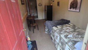 A compact room with a bed, a desk, and a chair. There is a red door, a ceiling light, and a wall-mounted picture of a moose.