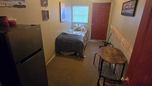 A compact room with a bed, a small table, a refrigerator, and a ceiling fan.