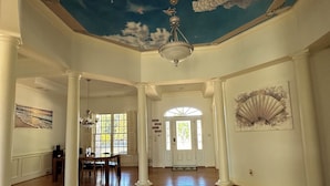A spacious room with wooden flooring, a chandelier, and a ceiling painted with clouds and a blue sky.