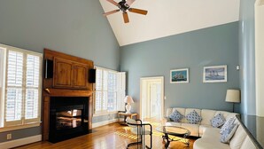 A spacious living room with hardwood floors, a fireplace, a ceiling fan, and a sofa with patterned cushions.