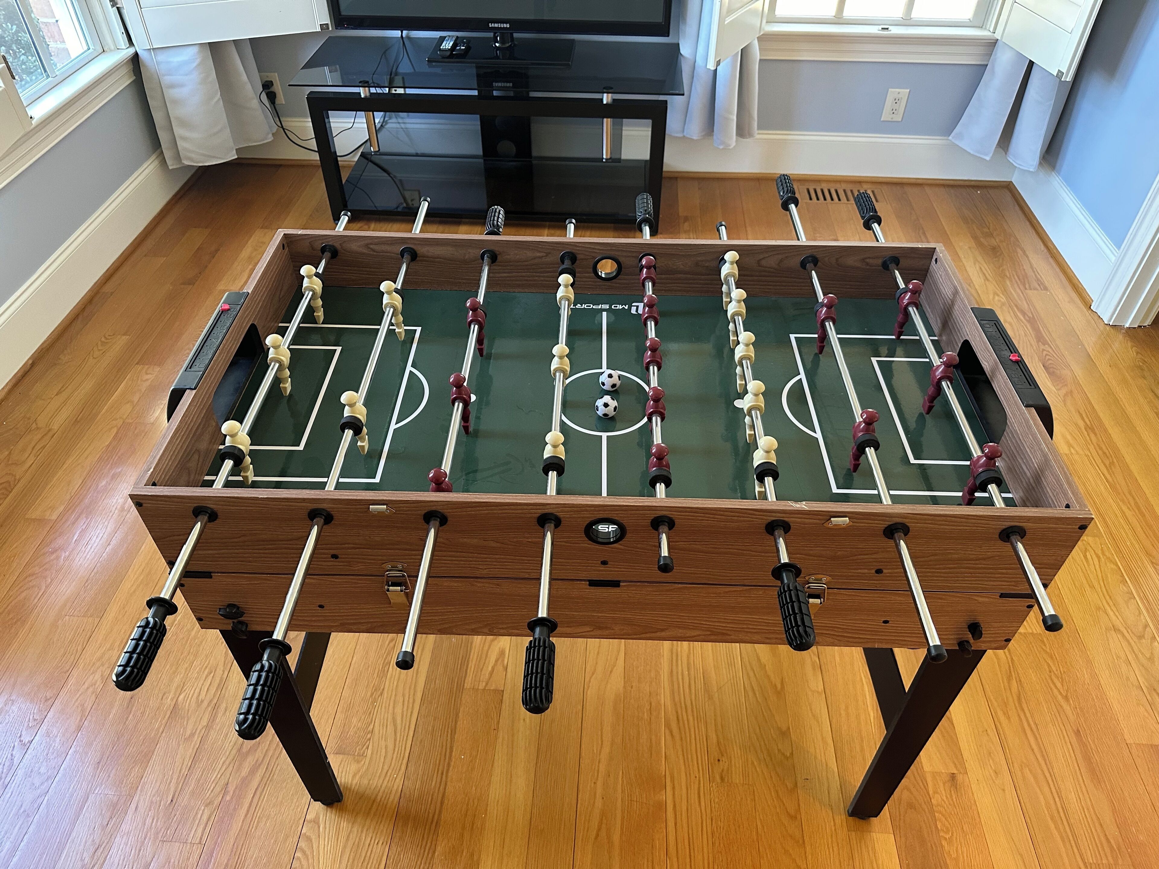 A foosball table in a room with wooden flooring, a television, and a window.