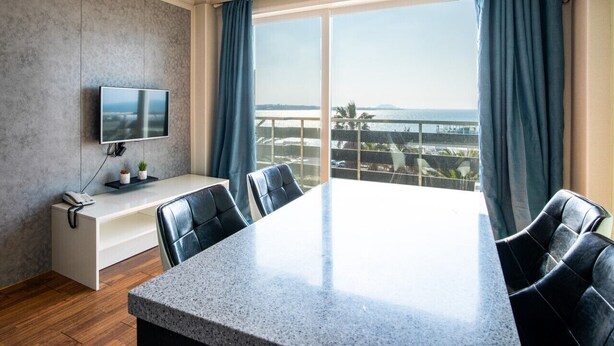 A modern hotel room with a large balcony, a flat-screen TV, a desk with a phone, and a conference table with chairs.