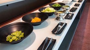 A buffet line with various food items including sliced kiwi, orange segments, and bowls of salad and soup.