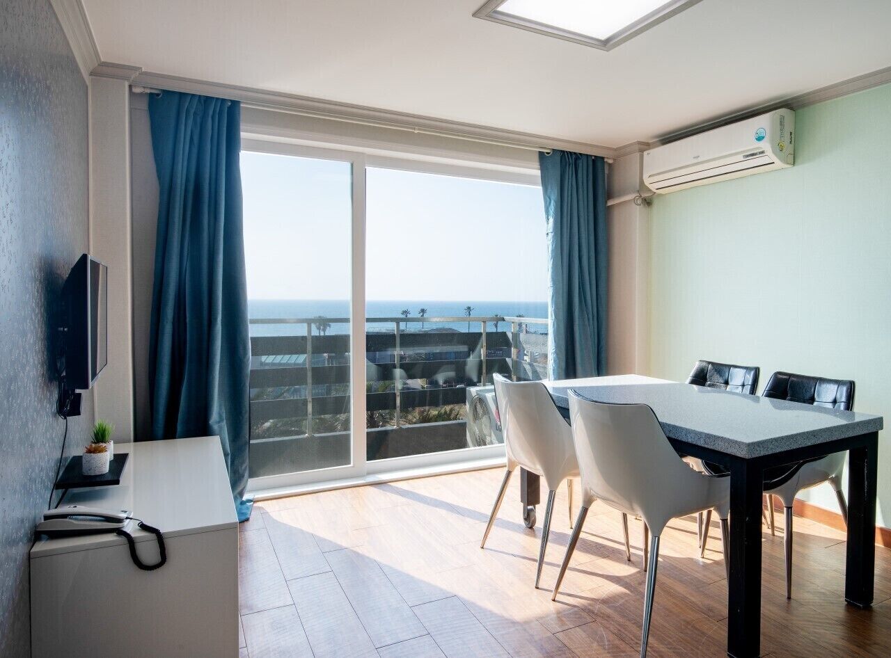 A modern room with a large sliding glass door leading to a balcony, a dining table with chairs, a small desk with a plant, and a mounted air conditioner.