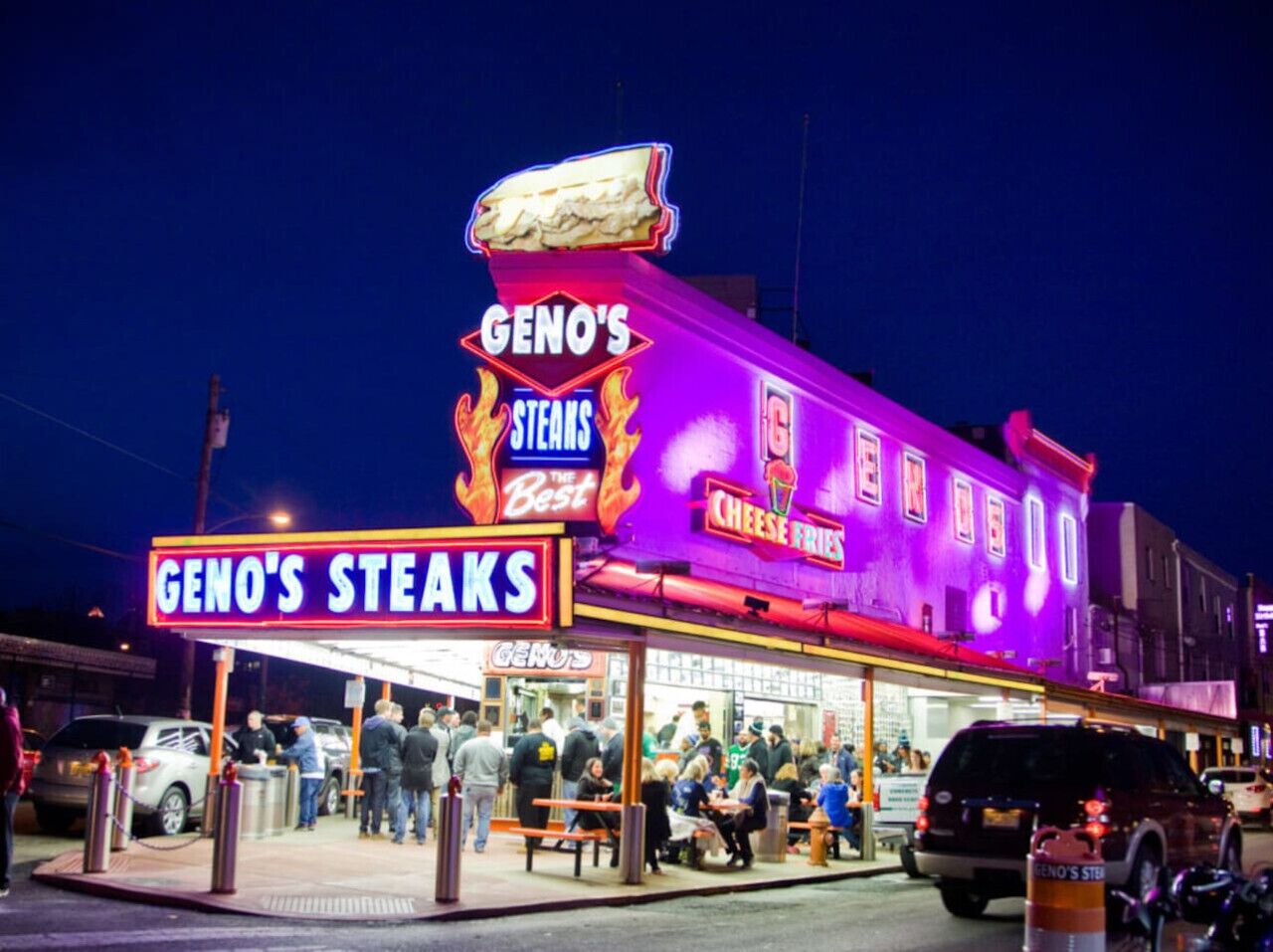 Um restaurante bem iluminado chamado 'GENO'S STEAKS' com letreiro de neon, mesas ao ar livre e um grupo de pessoas.