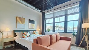 A modern bedroom with a bed, a pink sofa, a nightstand, and a window with a view. - Studio Apt Near Temple Unit with Free Parking for Extended Stays (Philadelphia)