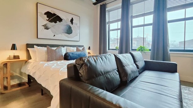 A modern bedroom with a bed, a black leather sofa, a bedside table, a lamp, and a window with curtains. - Cozy Philly Studio with Free Parking for Long Term Stays (Philadelphia)