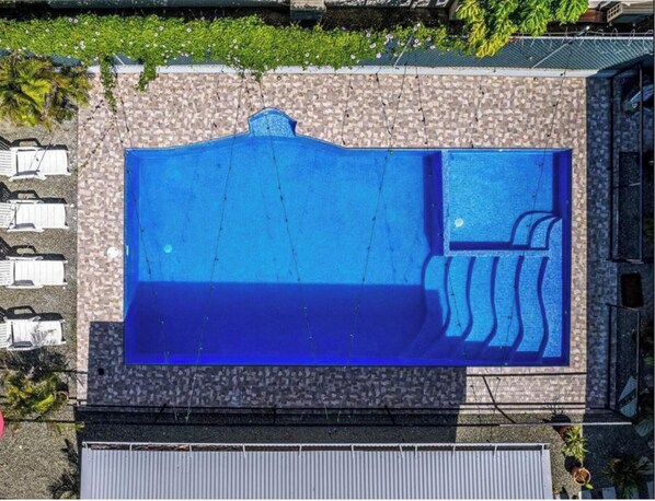 Vue aérienne d’une piscine bordée d’un mur en pierre, d’un petit jacuzzi attenant et d’une rangée de chaises longues blanches.