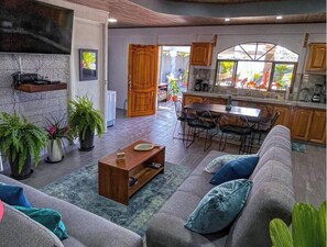 Uma sala de estar moderna com sofá, mesa de centro e plantas em vasos. Há uma área de refeições com mesa e cadeiras, e uma cozinha com armários e uma janela.