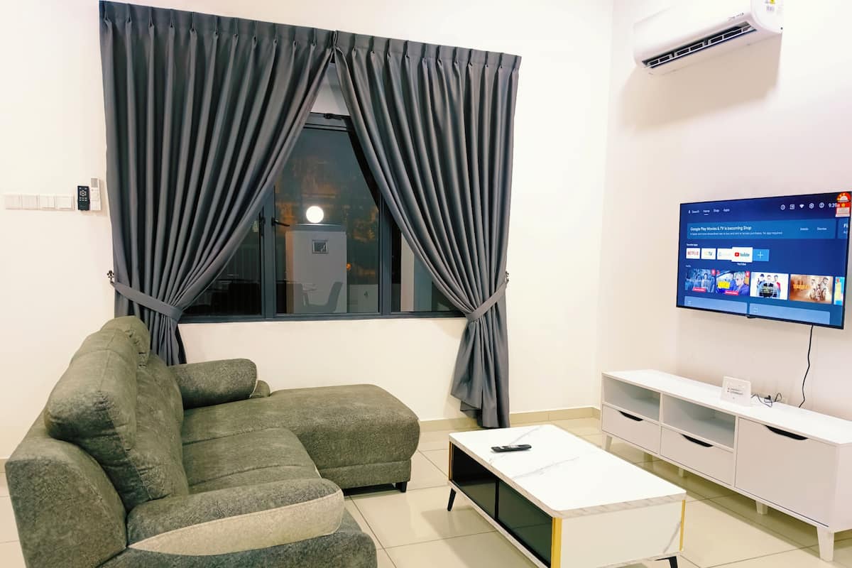 Una sala moderna con un sofá seccional, una mesa de centro blanca, un televisor de pantalla plana montado en la pared y una ventana con cortinas oscuras.