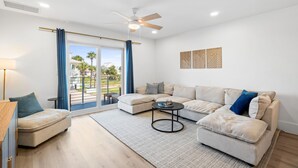 Una sala de estar moderna con un sofá, un sillón y una mesa de centro, un ventilador de techo y vistas a un espacio exterior a través de amplios ventanales.