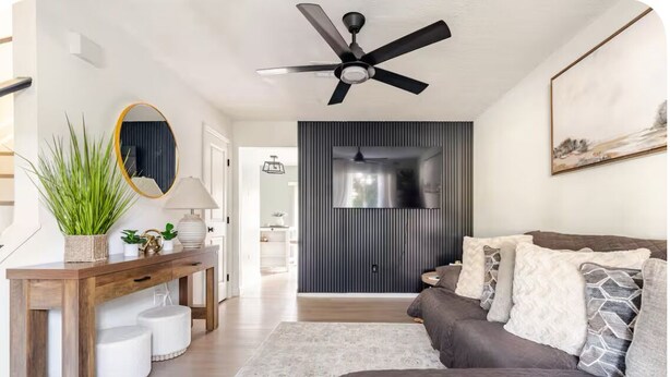 A modern living room with a ceiling fan, a sofa, a TV mounted on a wall, and a wooden console table with decorative items. - Charming 2-bedroom townhome in Winter Springs. (Winter Springs)