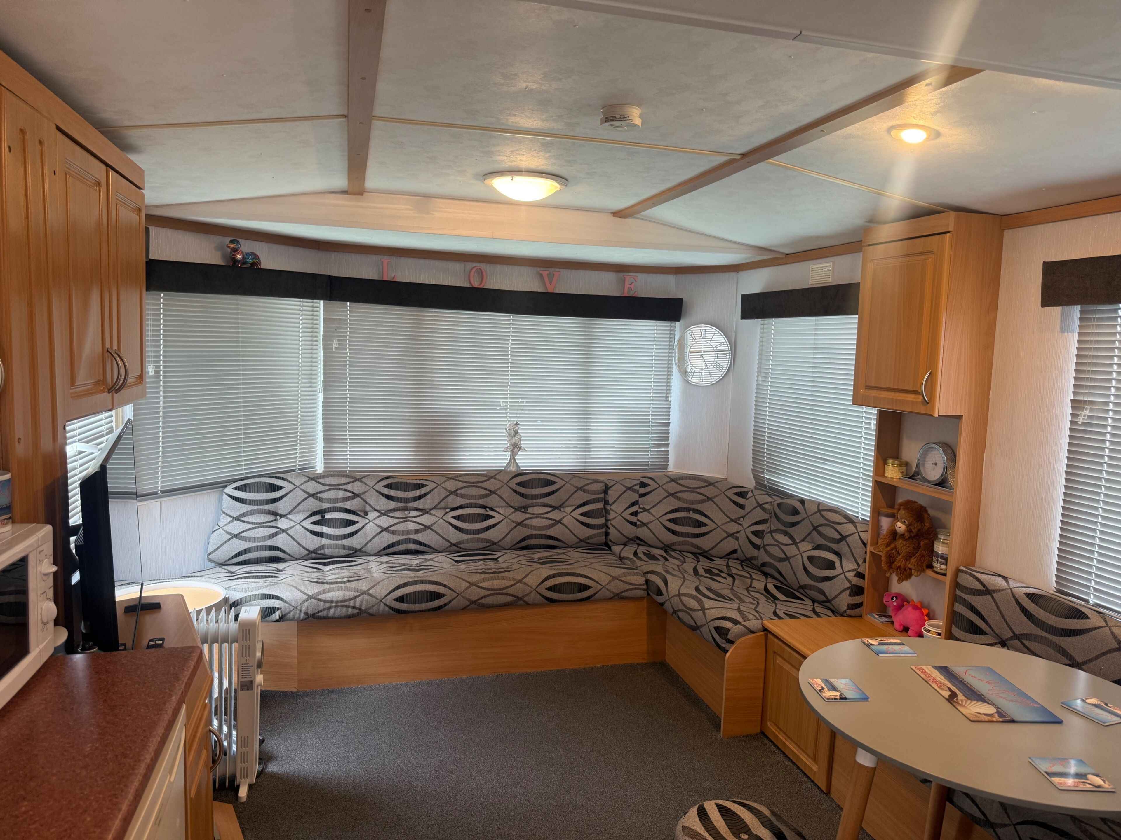 A compact RV interior with a kitchenette, a seating area with patterned cushions, a dining table, and a clock on the wall.