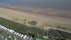 An aerial view of a coastal area with a sandy beach, a dense forested area, and a row of white buildings with green roofs.