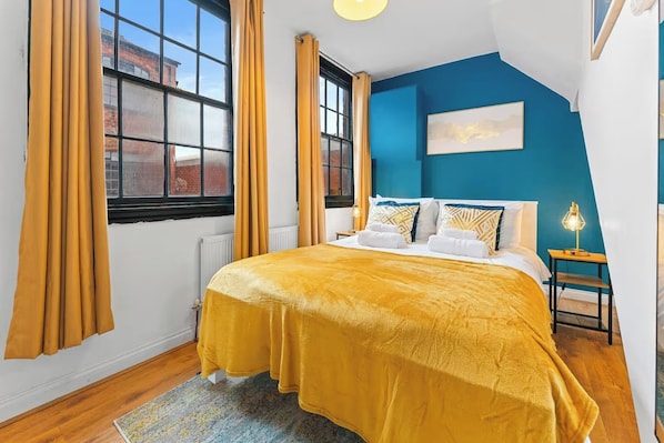 A bedroom with a large bed, a window with curtains, a blue accent wall, and a small bedside table with a lamp.